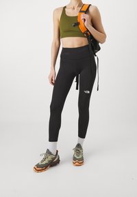 Svarta leggings med hög midja, tillsammans med en olivgrön sport-bh. En svart ryggsäck med orange rem fulländar träningsoutfiten.