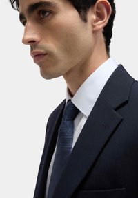 Granatowy garnitur o dopasowanym kroju, biała koszula wizytowa i strukturalny niebieski krawat. Wyróżniające się szczegóły to szpiczasty klap i kieszonka na poszetkę.