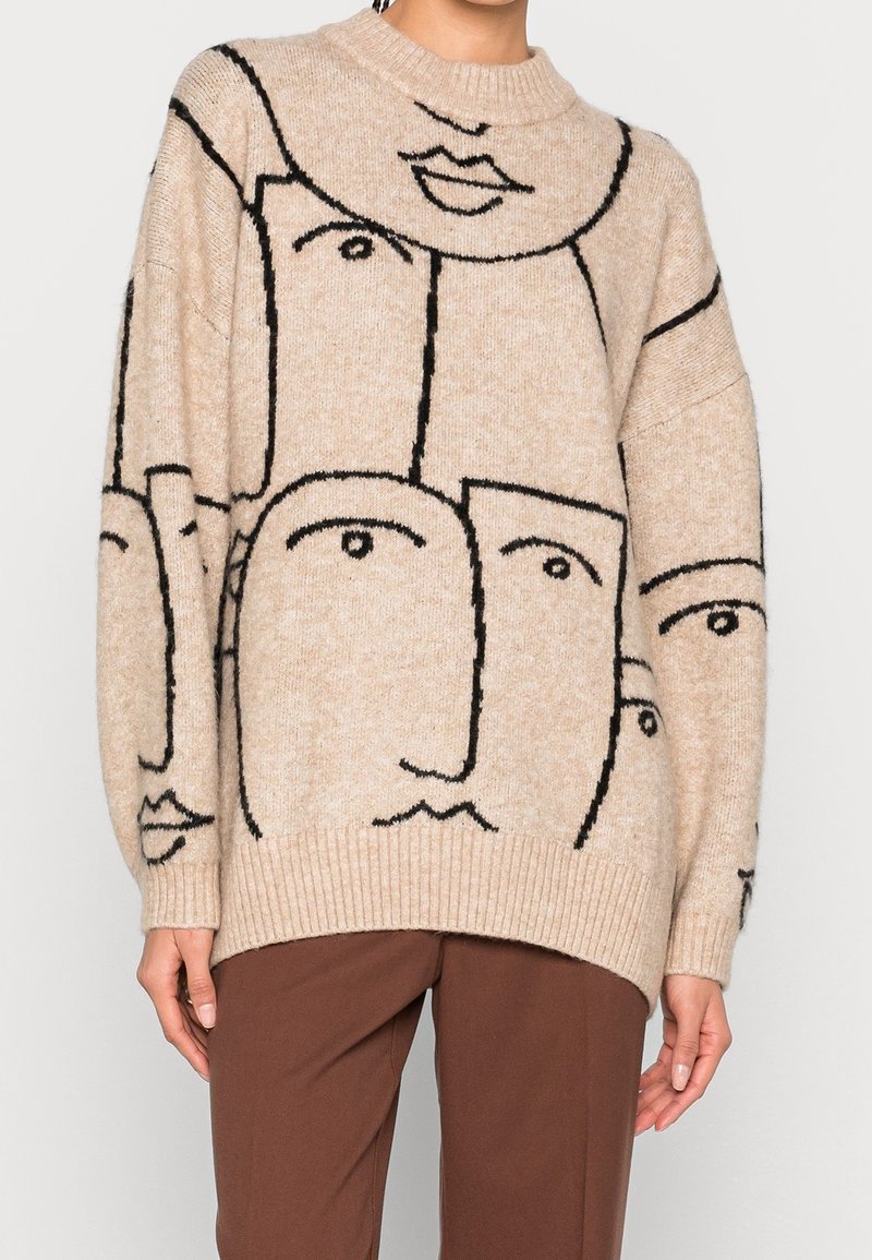 Överdimensionerad beige tröja med svarta ansiktsmotiv, rund halsringning, ribbade ärmmuddar och nederkant. Mjuk stickad textur, avslappnad silhuett.