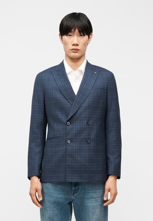 Ung mand iført en blå ternet dobbeltradet blazer over en hvid skjorte og blå jeans, stående foran en ensfarvet baggrund.