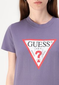 Lilla bomulls T-skjorte med et stort hvitt og rødt trekantet merke med merkevarenavnet "GUESS," detaljerte designelementer og et spørsmålstegn.