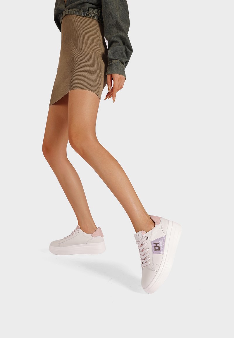 Sneakers bianchi con accenti rosa sul tallone e un logo decorativo sul lato. Il design include una chiusura con lacci e una suola spessa bianca.