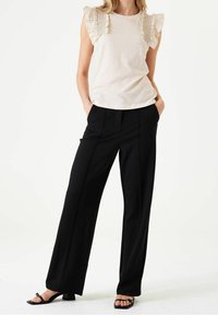 Donna con un top senza maniche beige con spalline arricciate, pantaloni neri a gamba larga e sandali neri con le dita scoperte, in piedi con le mani in tasca.