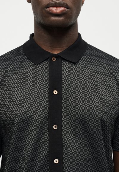 Άνδρας που φοράει πουκάμισο με κουμπιά, μαύρο και γκρι με σχέδιο, με μονόχρωμο μαύρο γιακά και καφέ κουμπιά, εμφανιζόμενος από τους ώμους έως τα χείλη.