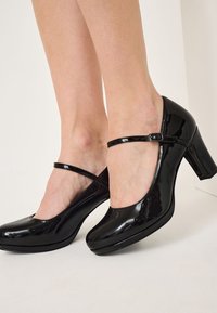 Kvinna som bär svarta lackläder Mary Jane-hälar med en tjock klack och spännband, stående på ett vitt golv mot en ljus vägg.