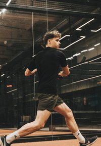 Jonge man in zwart T-shirt en korte broek doet een uitval om een squashbal te slaan op een binnenbaan met glazen wanden.