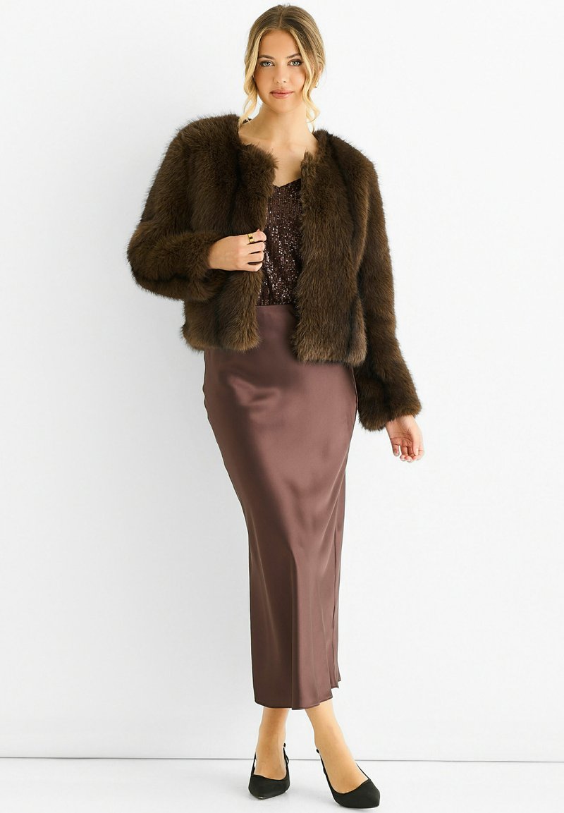 Woman wearing a dark brown faux fur jacket over a sequined top, paired with a satin midi skirt and black high-heeled shoes.