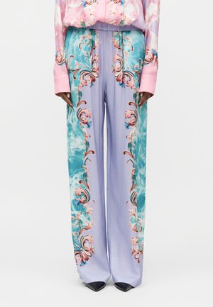 Mujer que lleva pantalones anchos de talle alto con patrones florales barrocos en azul, lavanda y rosa, y zapatos negros puntiagudos.