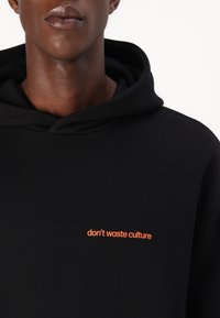 Schwarzer Hoodie mit einer Fronttasche und einer Kordelzugkapuze, mit orangefarbenem Text "don't waste culture" auf der Brust gestickt.