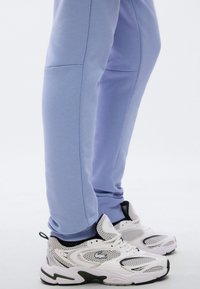 Ανοιχτό μπλε παντελόνια τζόγκινγκ με λεία, απαλή υφή και εφαρμοστή σχεδίαση, σε συνδυασμό με λευκά αθλητικά παπούτσια με λεπτομέρειες από δίχτυ και μαύρα διακοσμητικά στοιχεία.