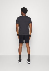 Schwarzes, kurzes T-Shirt mit kurzen Ärmeln, kombiniert mit schwarzen Shorts. Das Modell steht mit dem Rücken zur Kamera und betont das schlichte Design und die einfarbigen Farben.