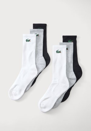 Deux ensembles de trois paires de chaussettes côtelées blanches, grises et noires avec un logo crocodile vert sur chaque bord-côte sur fond clair.