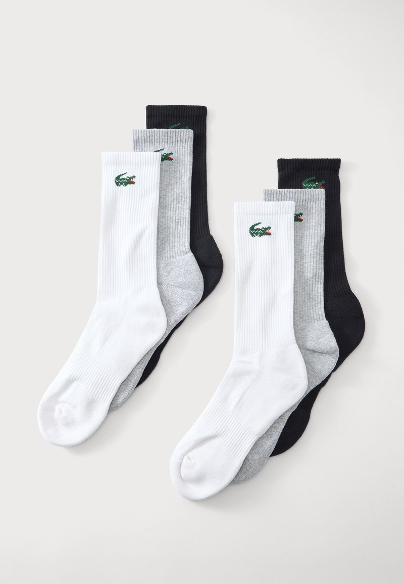 Deux ensembles de trois paires de chaussettes côtelées blanches, grises et noires avec un logo crocodile vert sur chaque bord-côte sur fond clair.