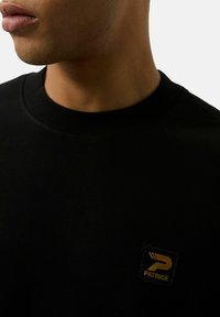 Felpa nera con colletto rotondo, realizzata in tessuto liscio. Presenta una toppa quadrata con testo e logo dorato sulla zona sinistra del petto.