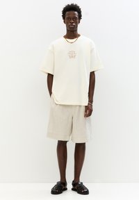 Cream short-sleeve t-shirt with central brown graphic design, paired with beige shorts and black strappy sandals. Minimalist style.