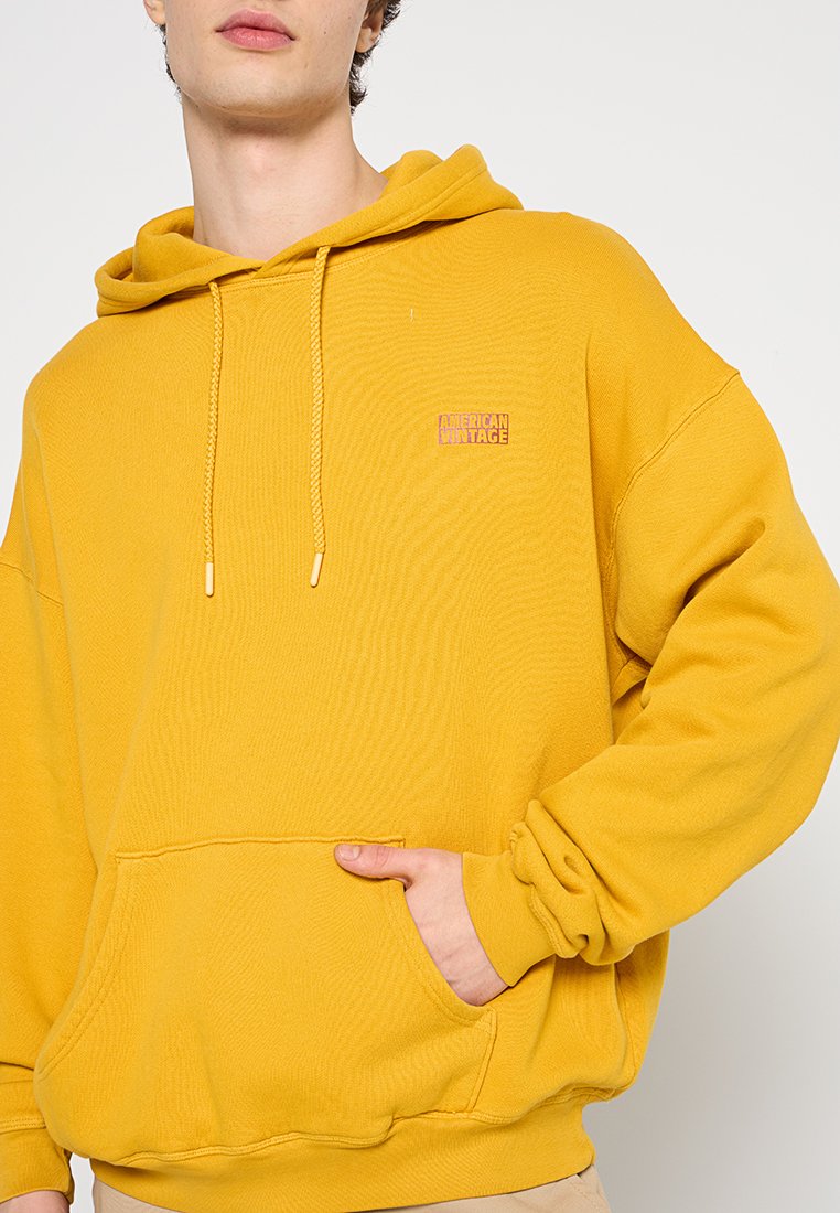 Geel katoenen hoodie met een kangoeroezak, trekkoordcapuchon en een klein logo-embleem op de borst. Heeft ribbels aan de mouwen en onderkant.