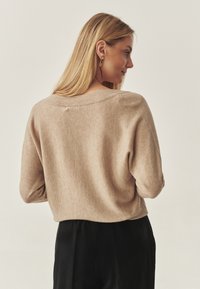 Pull beige en maille avec une coupe décontractée, encolure large et poignets côtelés, associé à un pantalon foncé, vu de dos.