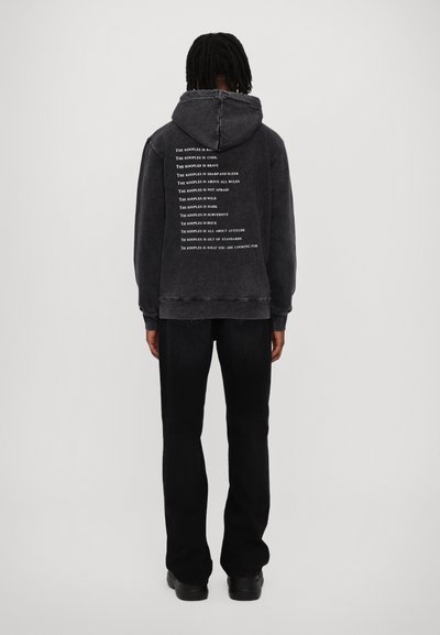 Person with dark braided hair wears faded black hoodie with white text on back and black pants, standing against plain light background.