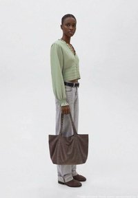Brązowa torebka z zamszu z dwiema długimi rączkami i ćwiekowymi akcentami, zestawiona z jasnozieloną bluzką i jasnymi dżinsami, na jednolitym tle.