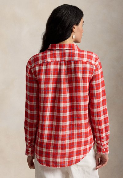 Kvinne med langt mørkt hår iført rød og hvit rutete skjorte med lange ermer, vist bakfra mot en ensfarget nøytral bakgrunn.