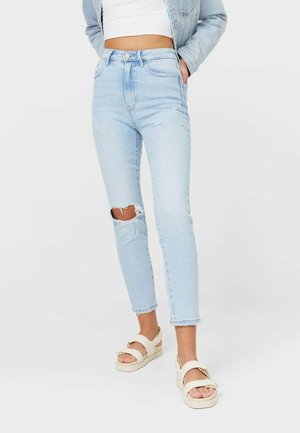 Femme portant un jean skinny bleu clair déchiré, un crop top blanc, une veste en jean claire et des sandales compensées beiges, debout devant un fond uni.