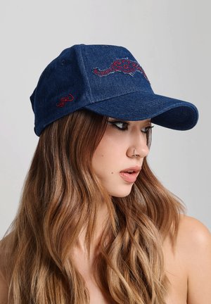 Jeune femme aux longs cheveux ondulés portant une casquette de baseball en jean bleu avec une broderie rouge, regardant sur le côté sur un fond uni.