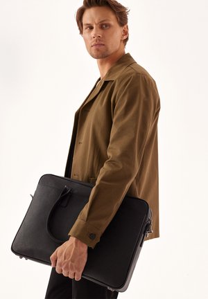 Maletín de cuero negro con un asa superior, forma rectangular, textura suave y cierre de cremallera en la parte frontal, sostenido por una persona que lleva una chaqueta marrón.