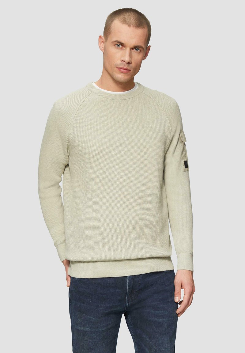 Jasno beżowy sweter z dzianiny o okrągłym dekolcie i raglanowymi rękawami. Ma teksturowaną tkaninę oraz małą kieszonkę na rękawie.