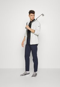 Stroje golfowe Nike, składające się z jasnoszarej kurtki, czarnego polo, granatowych spodni i szarych butów. Kurtka zapinana na zamek z czarnymi akcentami.