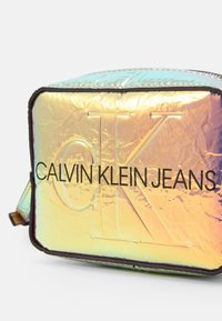 Irisfärgad rektangulär väska med texturerad yta, med texten "CALVIN KLEIN JEANS" i svart. Försedd med dragkedja och justerbar rem.