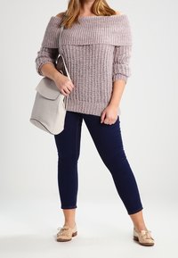 Hellrosa, schulterfreier Strickpullover mit geripptem Muster, kombiniert mit dunklen Skinny-Jeans und beigen Slippern, während eine hellgraue Tasche gehalten wird.