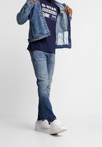 Denimjacka i ljusblått med tryckknappar. Marinblå t-shirt med vit text och jeans i medelmörk tvätt. Vita sneakers.