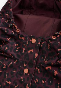 Lähivõte burgundiapunase jakist, millel on musta ja oranži leopardimustriga detailid, oranžid pressnööbid ja reguleeritavad kapuutsiköied.