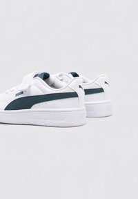 Baskets Puma blanches avec une bande d'accent verte, présentant une surface texturée, un col rembourré et une semelle en caoutchouc plate. Logo visible sur le côté.