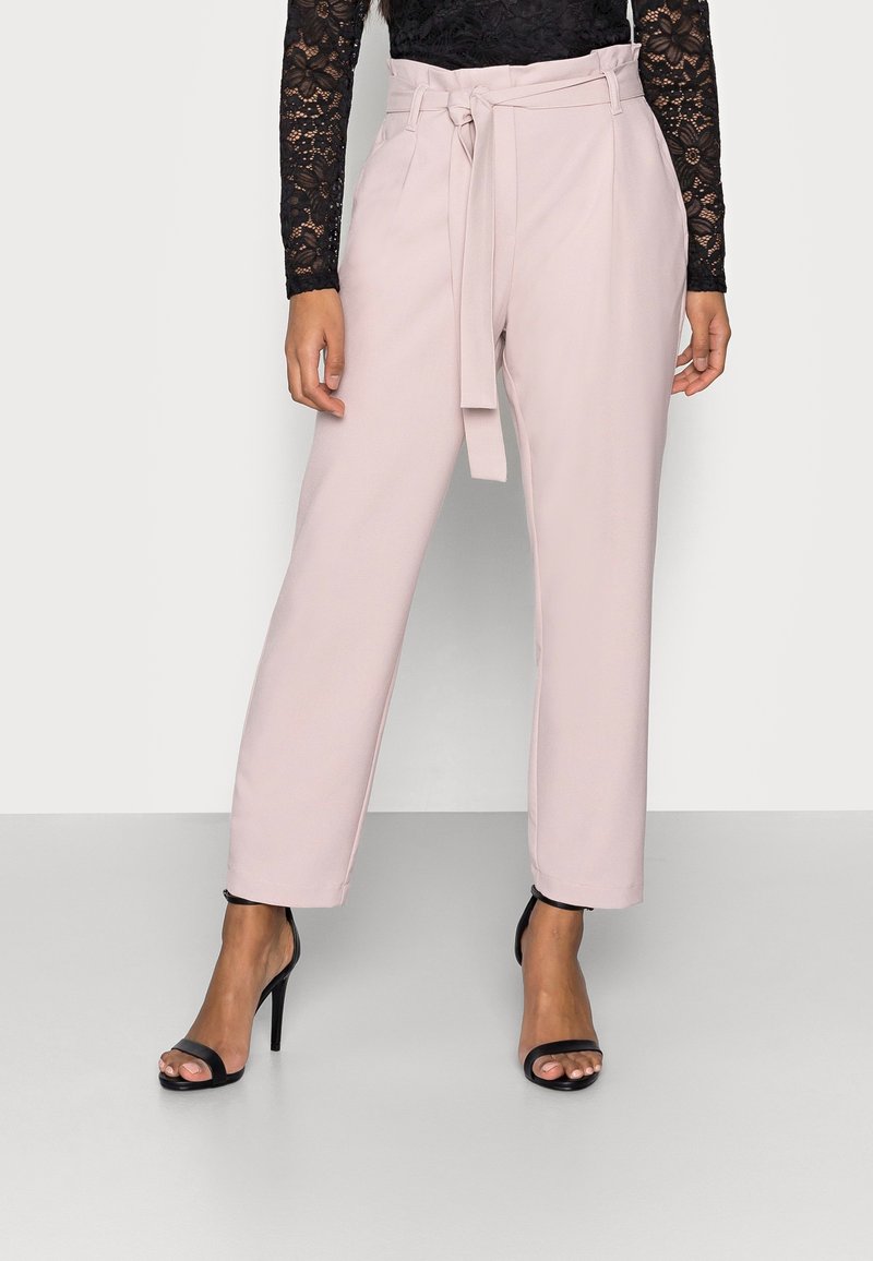 Pantalon rose clair sur mesure avec une taille haute, une ceinture à nouer et des jambes larges, associé à des talons noirs à lanières et un haut à manches longues en dentelle noire.