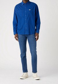 Blå långärmad skjorta med knäppning, två framfickor, matchad med slim-fit blå jeans och vita sneakers, mot en ljus bakgrund.