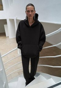 Felpa nera oversize con una grande tasca frontale e cappuccio con cordini, abbinata a pantaloni scuri sartoriali. Presenti lunghi orecchini di grande impatto.