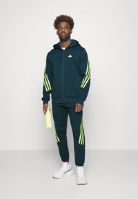 Tamno tirkizna majica sa zatvaračem i neonsko zelene pruge na rukavima, odgovarajuće trenerke i bijele tenisice. Drži svijetlozelenu bocu za vodu.