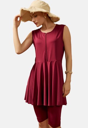 Donna con vestito bordeaux senza maniche con zip frontale, shorts abbinati fino al ginocchio, cappello beige a tesa larga, orecchini a conchiglia e bracciale.