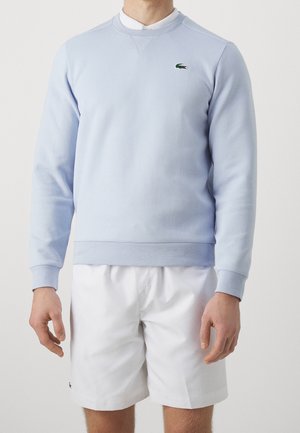 Mann trägt hellblauen Sweatshirt mit grünem Krokodil-Logo und weißen Shorts vor einfarbigem Hintergrund, steht mit entspannten Armen.