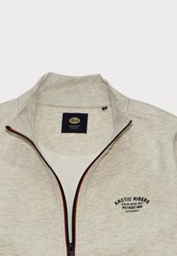 Světle šedá zipová bunda vyrobená z měkké tkaniny. Má vysoký límec, černý zip s červeným akcentem a logo "ARCTIC RIDERS" na prsou.