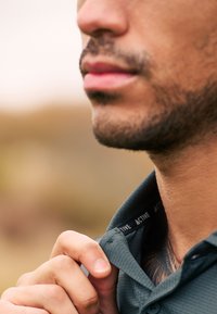 Polo shirt de sport gris foncé avec un tissu texturé, détail de col visible, et une étiquette portant "ACTIVE" près du cou. Le ton de peau et les traits du visage sont partiellement visibles.