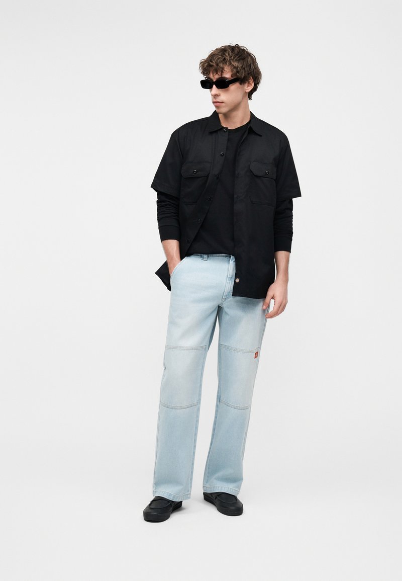 Junger Mann mit lockigem Haar, der schwarze Sonnenbrillen, geschichtete schwarze Hemden, hellblaue Jeans und schwarze Schuhe trägt und mit einer Hand in der Tasche steht.