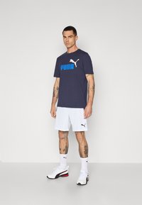 Navy t-shirt with blue "PUMA" logo, white shorts, black Puma logo, white sneakers with red accents, displayed against a plain background.