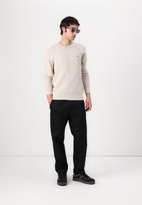 Pull beige clair à col rond avec un petit logo sur la poitrine, associé à un pantalon noir et des baskets noires. Des lunettes de soleil claires sont portées.