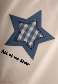 Zilā zvaigznes plāksteris ar rūtainu rakstu un melnu šuvi uz bēša auduma, zem tā izšūts teksts "Mēs visi esam zvaigzne".