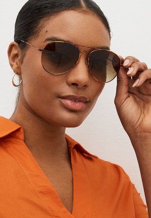 Sieviete valkā bruņurupuču čaumalas aviatoru saulesbrilles un apelsīnu krāsas krekla apkakli, vienu roku pielāgojot saulesbrilles.