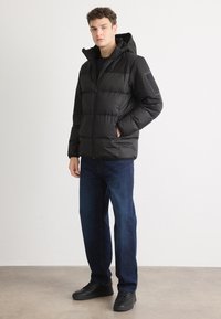Zwarte pufferjack met capuchon, ritssluiting en een gestructureerde stof. Dragen met blauwe jeans en zwarte sneakers, met zijzakken en een schouderpatch.
