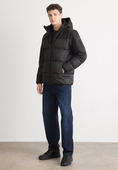 Doudoune noire avec capuche, fermeture éclair et tissu texturé. Portée avec un jean bleu et des baskets noires, dotée de poches latérales et d'un patch sur l'épaule.