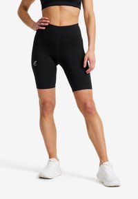 Kvinna iklädd svarta, tighta cykelbyxor och vita sneakers, stående med en hand vilande på sin höft mot en enfärgad bakgrund.